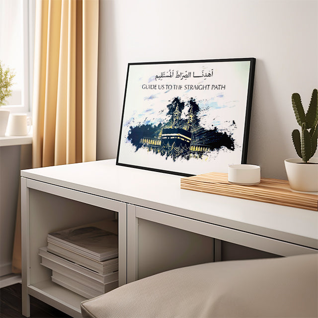 Framed artwork on a desk with a plant and books in a room.